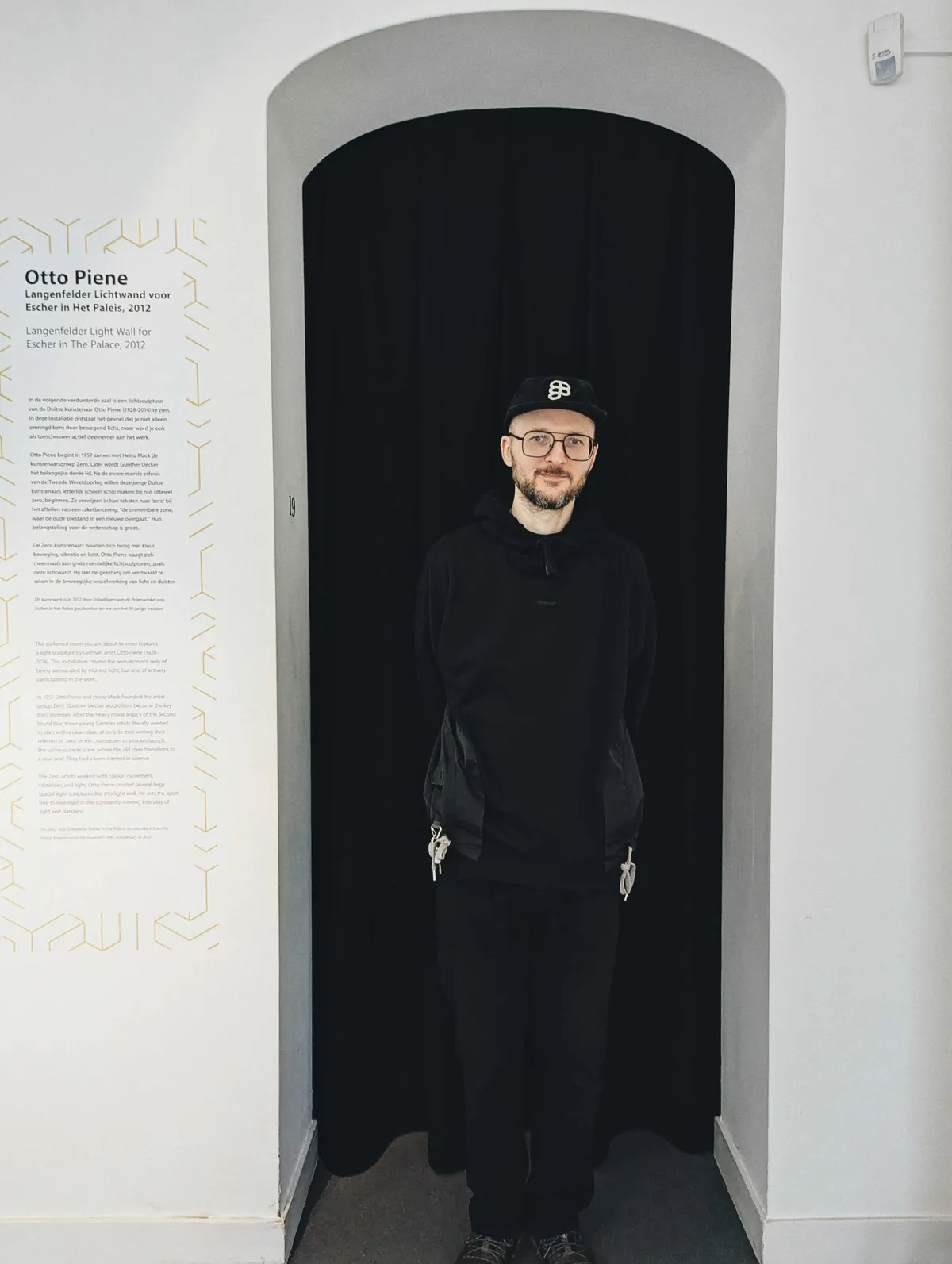 Daniel, wearing all black, standing in front of a black curtain with his hands behind his back.