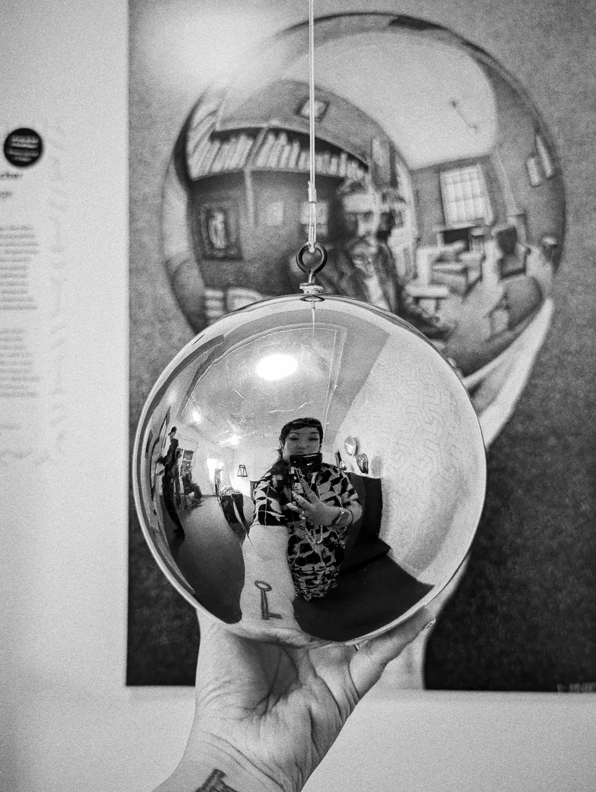 A black and white image of Peige holding a mirrored orb, imitating the image of Escher in the background.