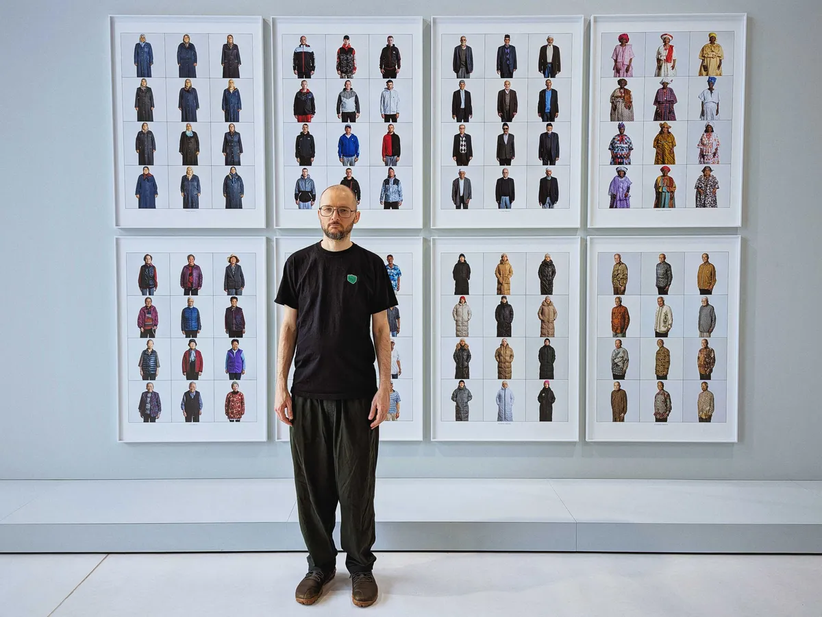 Daniel stands in front of a series of portraits. The portraits are grouped by ethnicity and depict people wearing similar clothing.