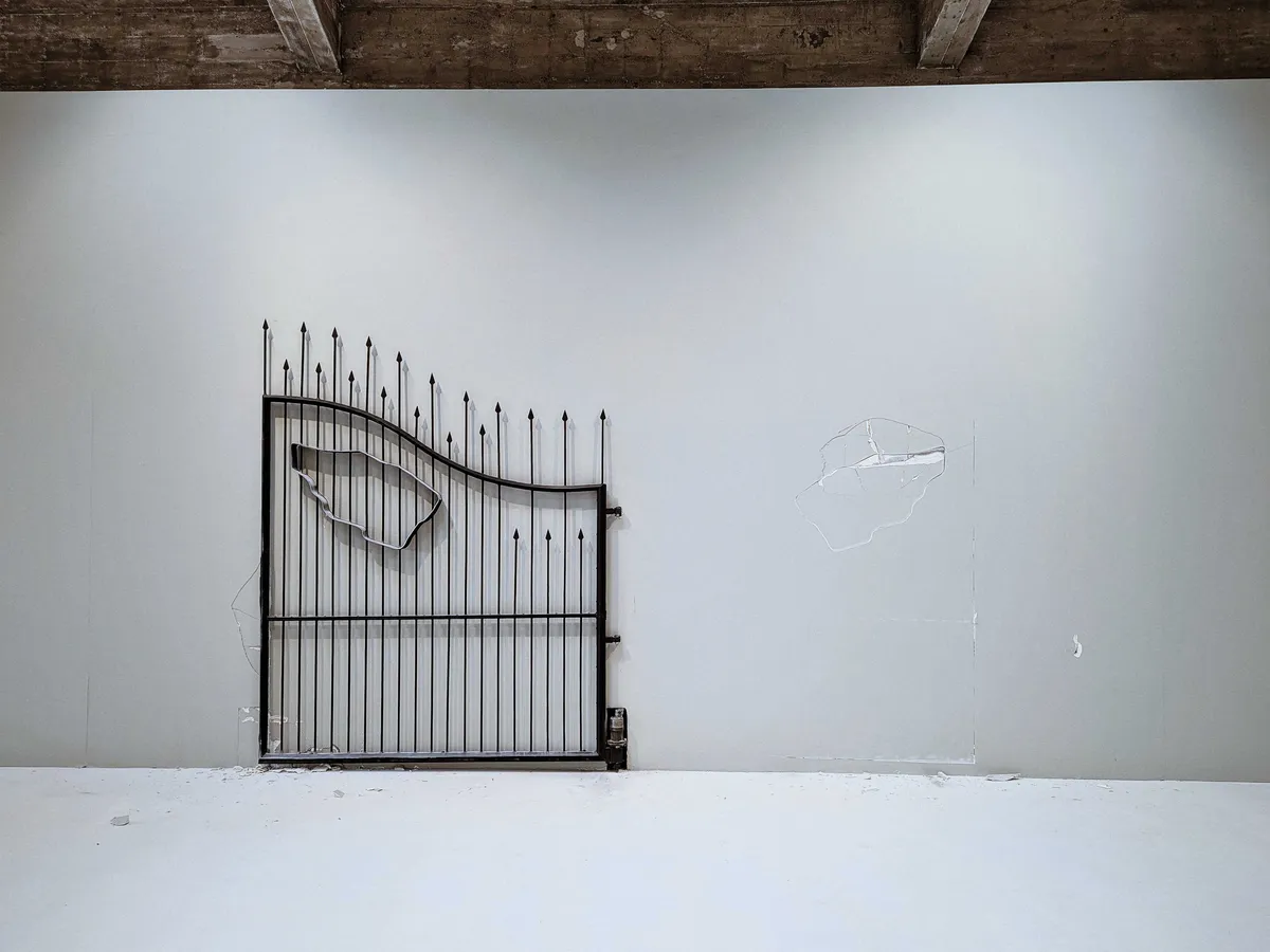 A single large, black metal gate with long spikes and the outline of a territory stands to the left of the wall. The right is a crack with the imprint of the outline.