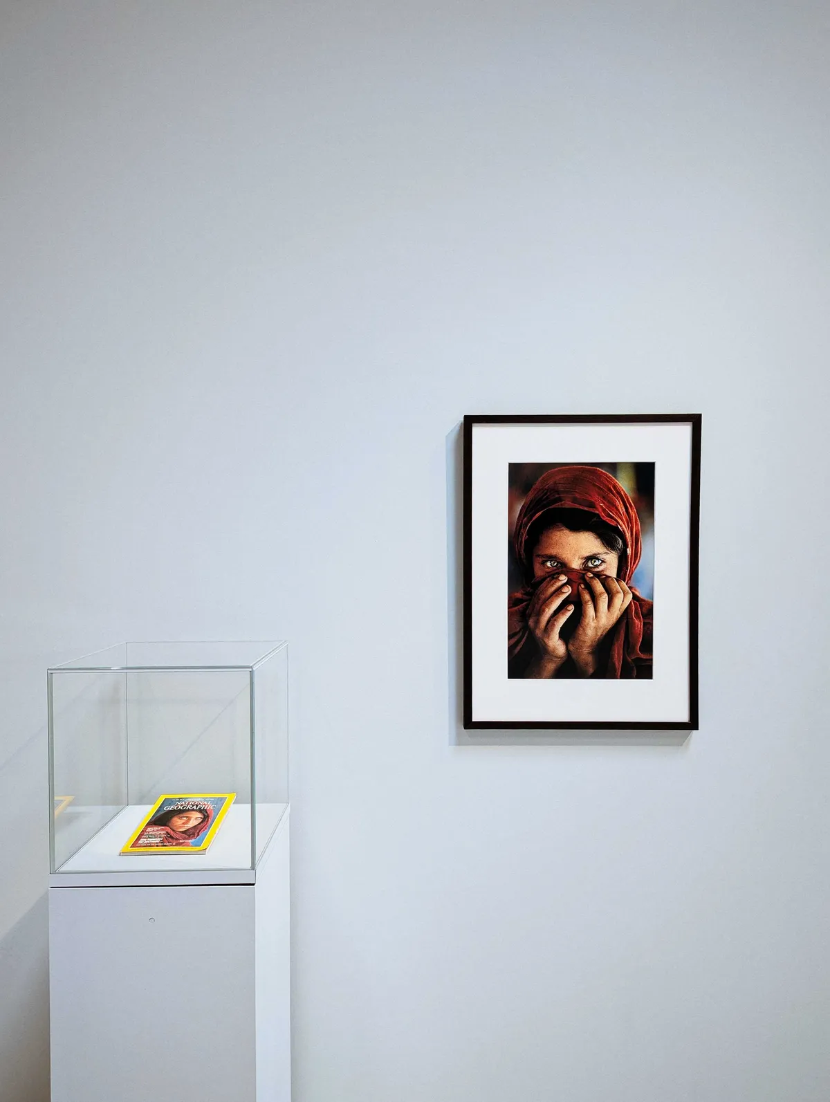 A copy of the infamous National Geographic issue of the Afghan girl is contained inside a glass case. On the wall is an image of the same girl, except she is covering her mouth with her hands and scarf. Her eyes looking bright and happy.
