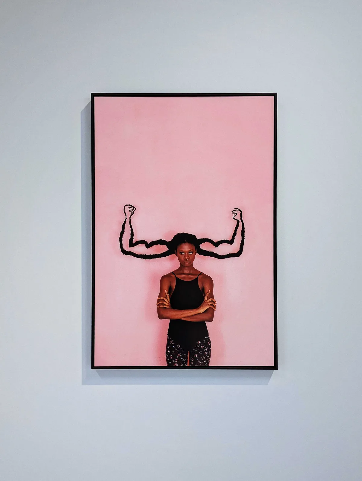 Portrait of a Black woman standing with her arms crossed in front of a pink background. Her hair has been woven into the shape of two muscular arms, sticking out from the sides of her head.