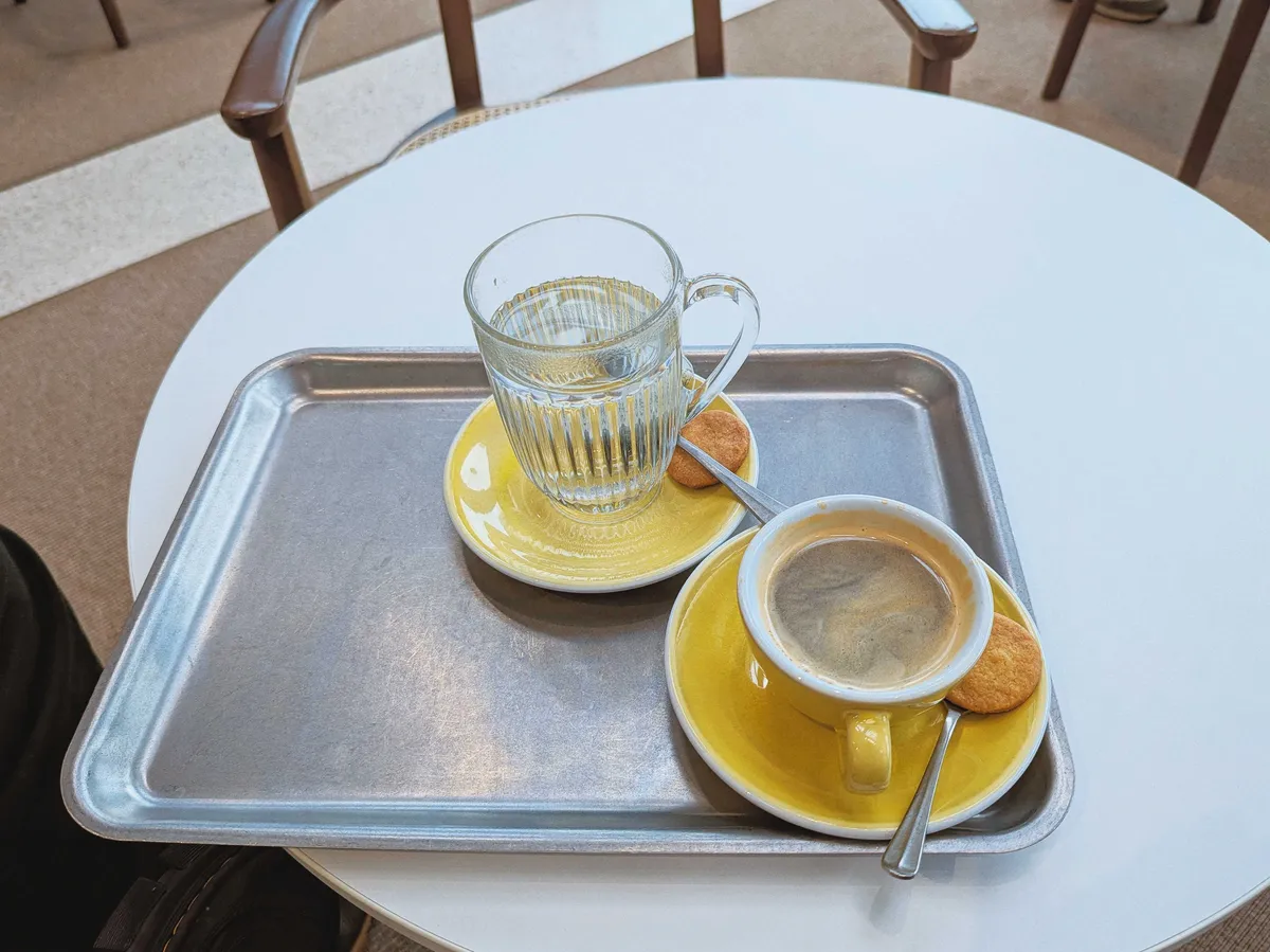 Coffee in a yellow ceramic cup and matching saucer with a small biscuit and spoon on the side. There is also a glass cup filled with water, also on top of a yellow saucer, with a biscuit and spoon. Both sit on a worn, silver tray.