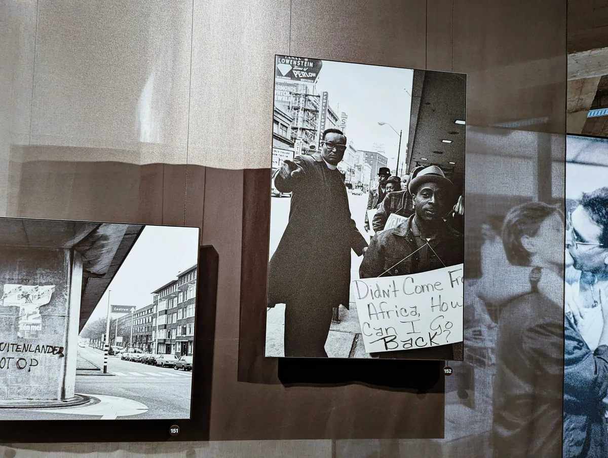 Photo exhibition, focusing on a black and white image of two Black men, one who has a sign around his neck reading, 