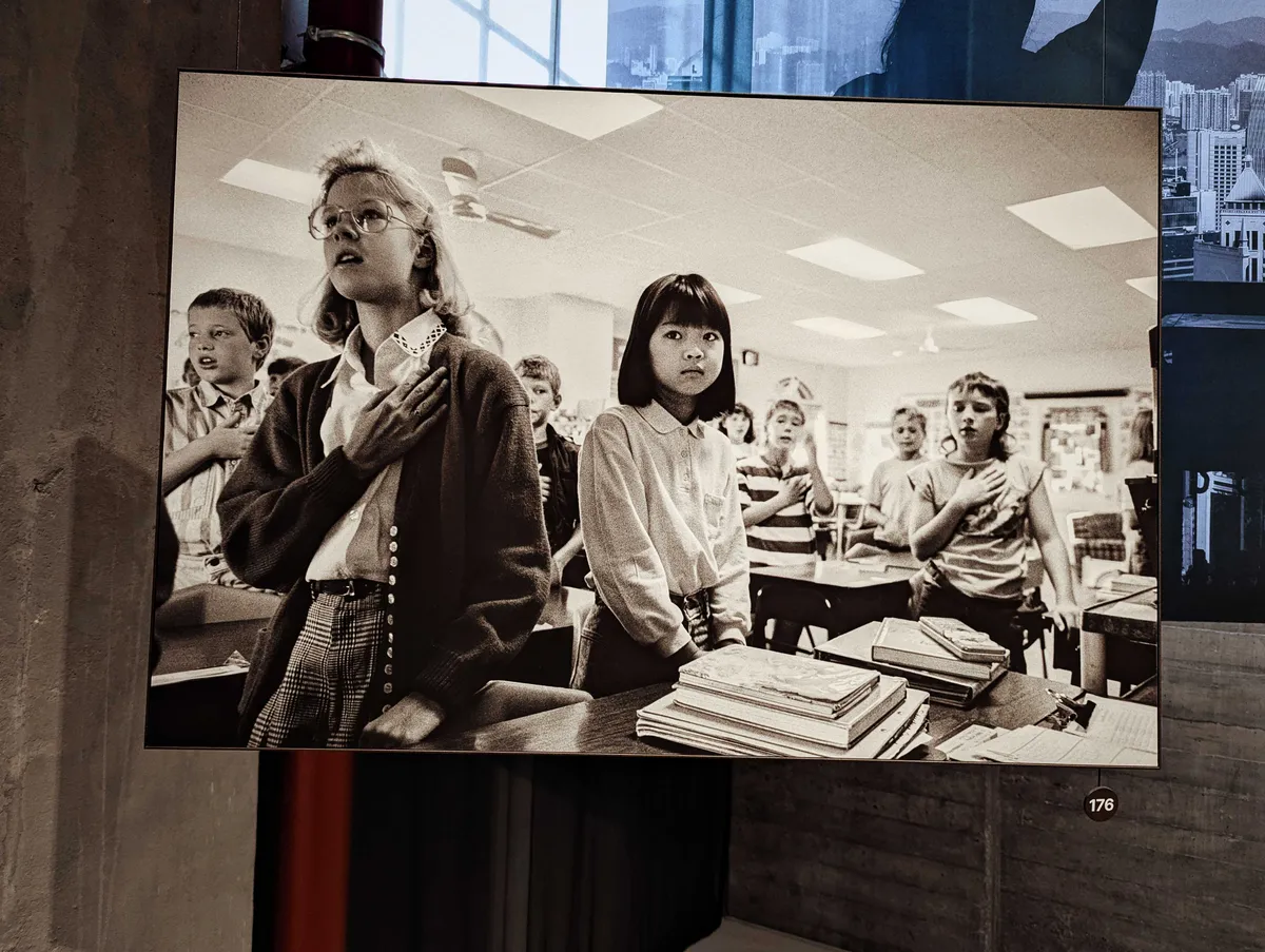 A large, black and white photo of white children from the 1980s, standing with their hands on their heart and reciting the pledge of allegiance. In the center is an Asian girl, glancing towards the side and looking nervous, with both hands down.