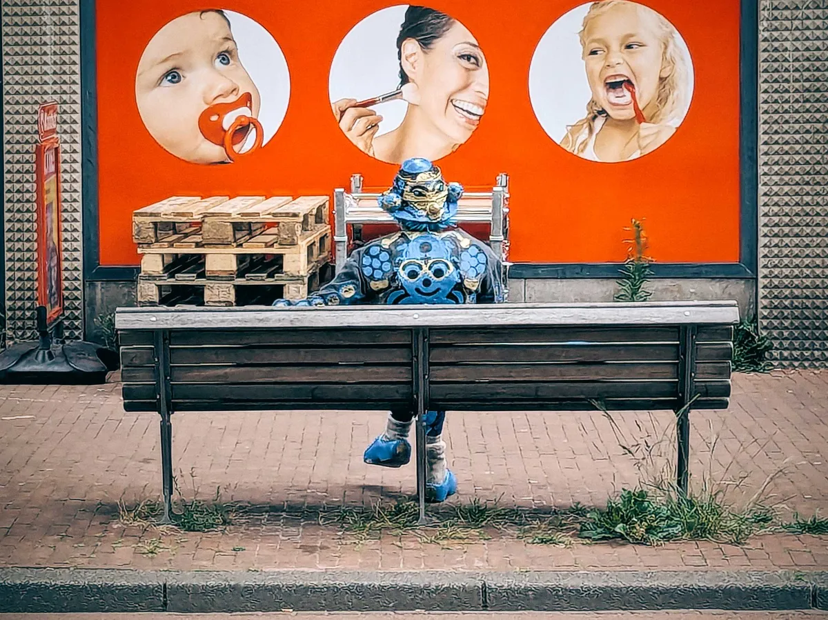 A person sits on a bench with their back facing the camera. They are wearing customized clothing painted in blue and gold, with a samurai helmet and a leather jacket with a smiling person painted on it.