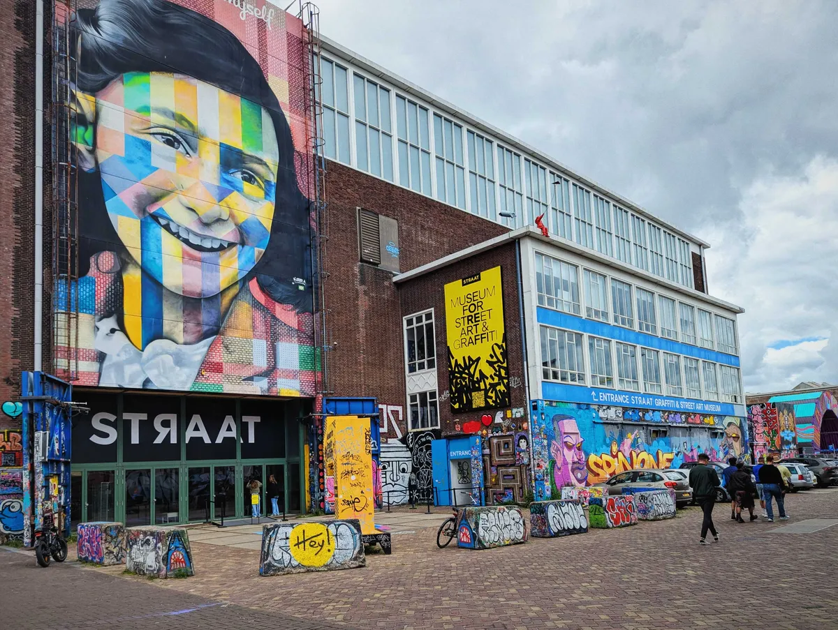 A brick warehouse building that is covered in street art, with signage indicating that its the STRAAT museum. Above the entrance is a large, stylized portrait of Anne Frank.