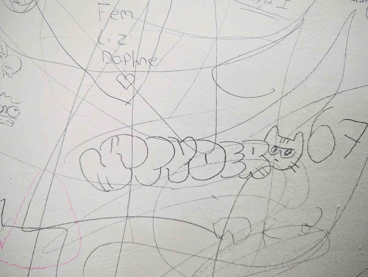Close-up of a white wall that has been drawn all over by various people in pencil. In focus is a tag by Peige that says, 