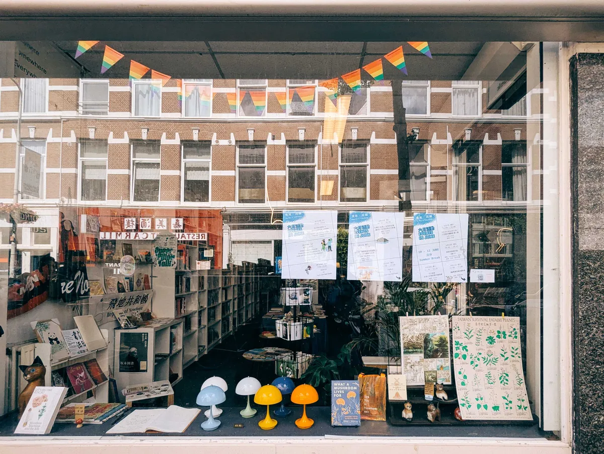The storefront window, with rainbow flag bunting hanging from the top, some colorful lamps displayed for sale, and lots of books and posters.