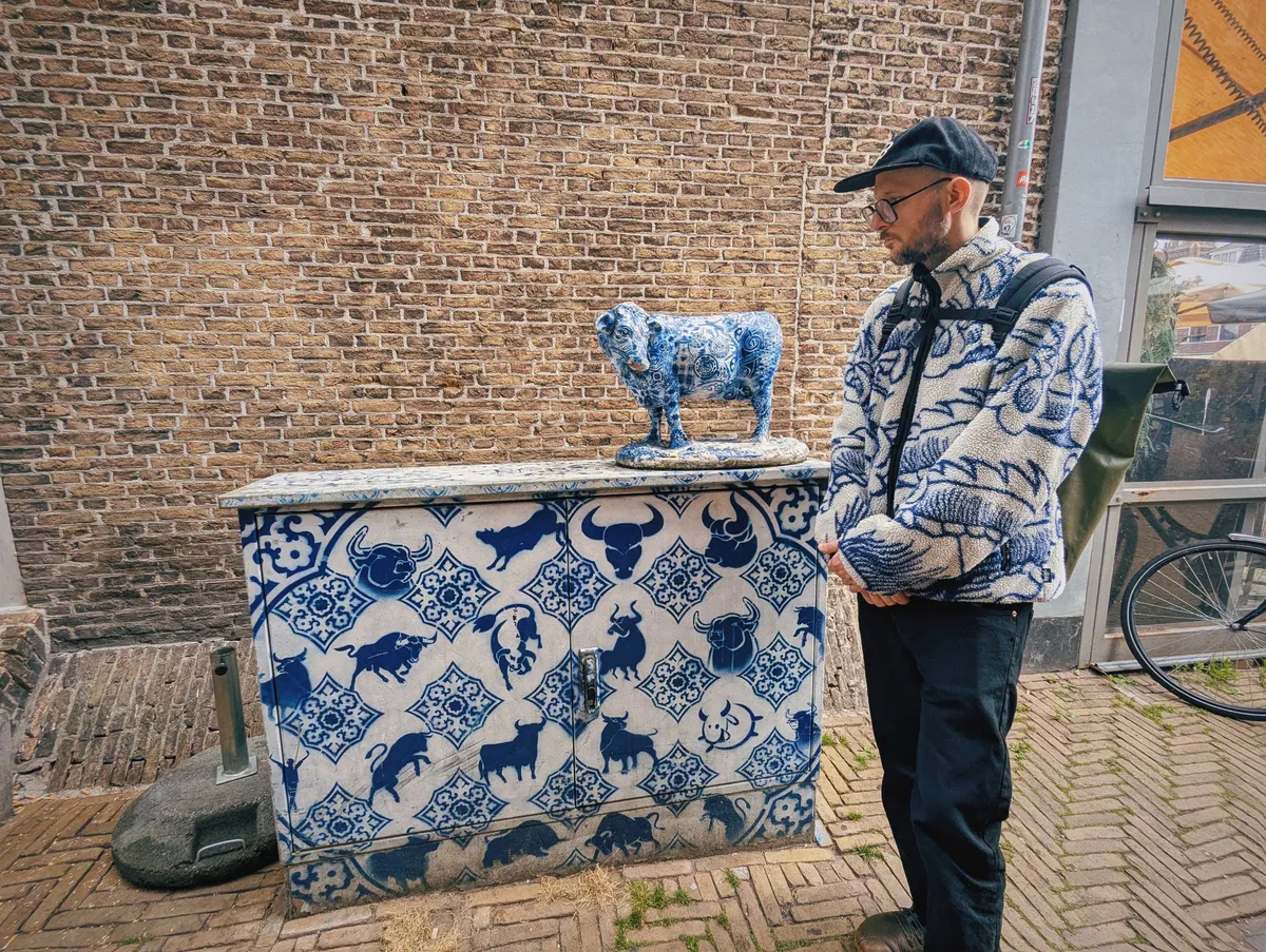 Daniel wearing a white fleece jacket with blue line art of a large dragon, stands next to a junction box that's been spray-painted white with blue Delftware inspired patterns and bulls. There's also a blue Delftware cow on top of the box.
