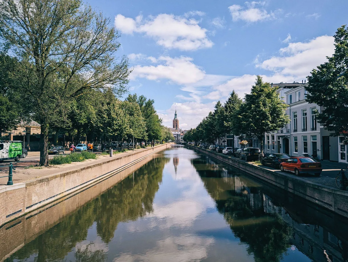 A scenic canal with dark green water. In the distance is the clock tower of Grote of Sint-Jacobskerk.