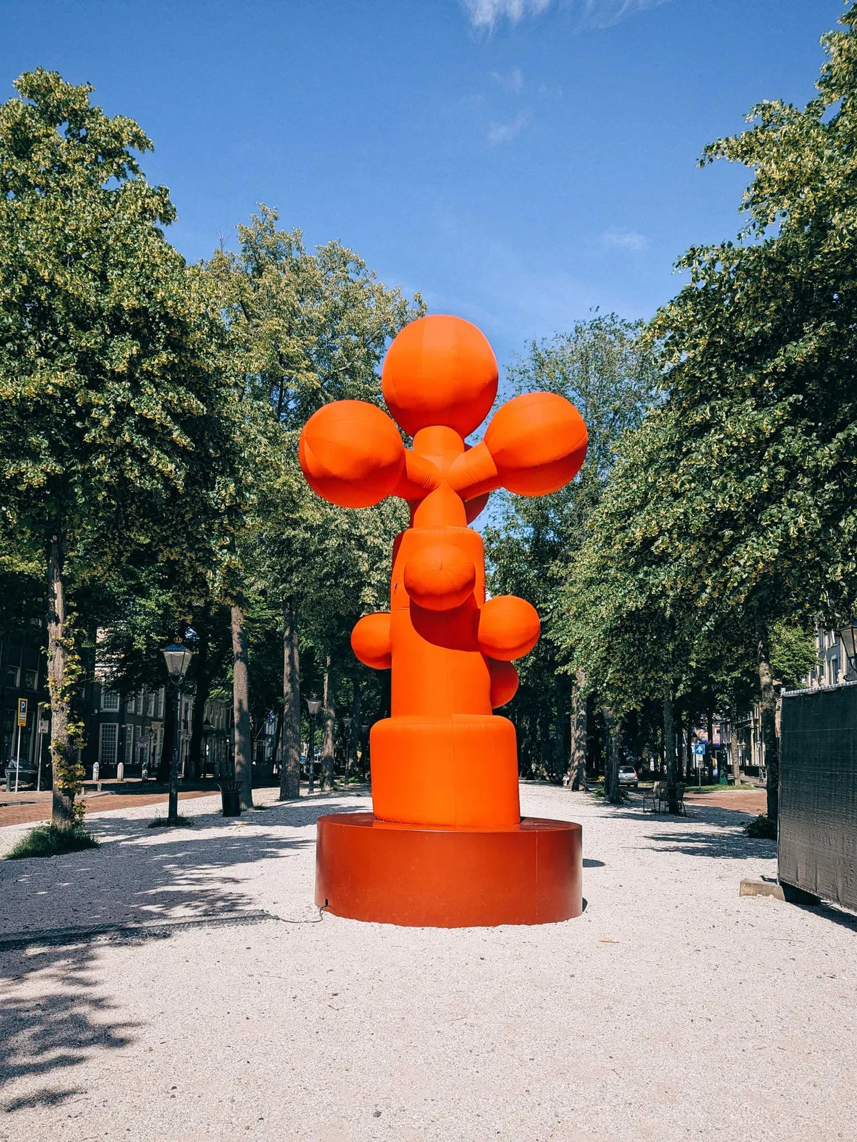 A bright orange inflatable gingko biloba-shaped sculpture stands in the center of a walkway through a park.