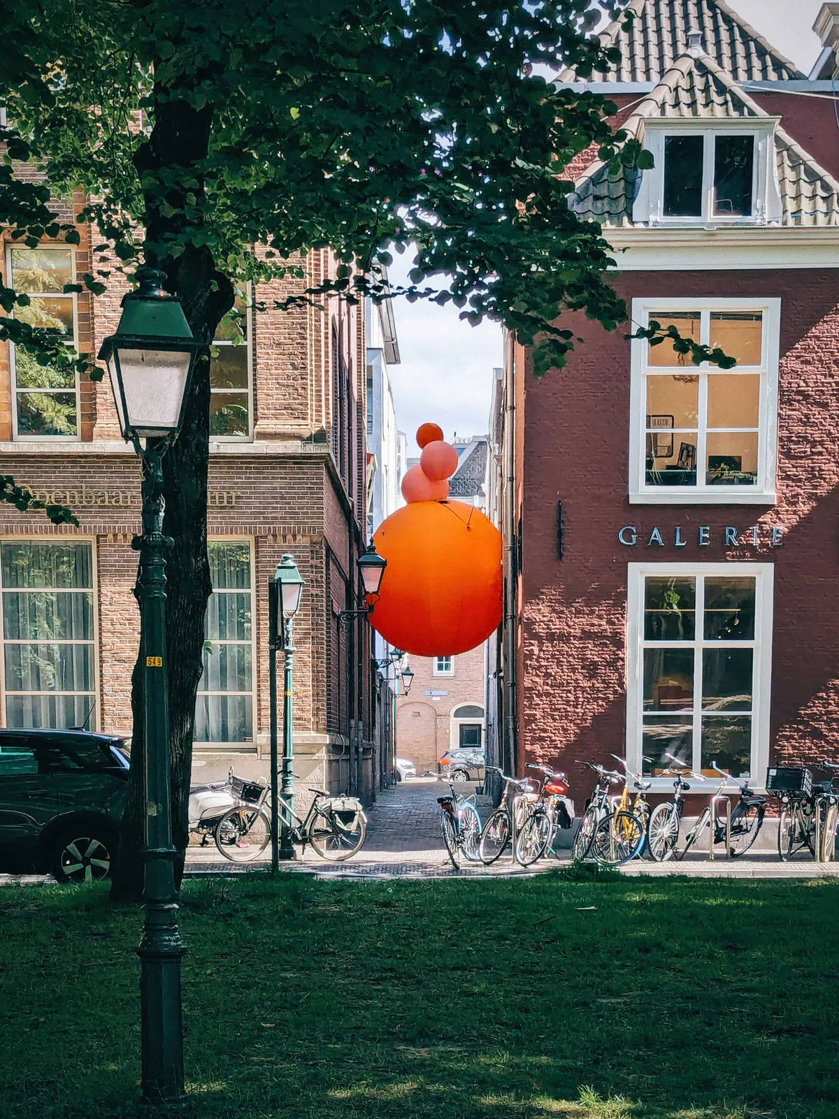 A large orange sphere with three smaller spheres on top are sandwiched between two brick buildings.