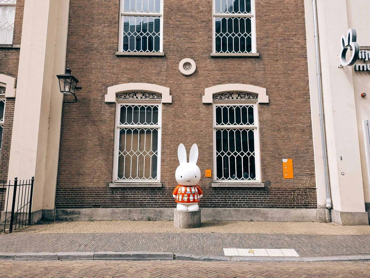 A statue of Miffy wearing a red shirt with other Miffys painted on it. It stands outside of a brick building with ornate windows.