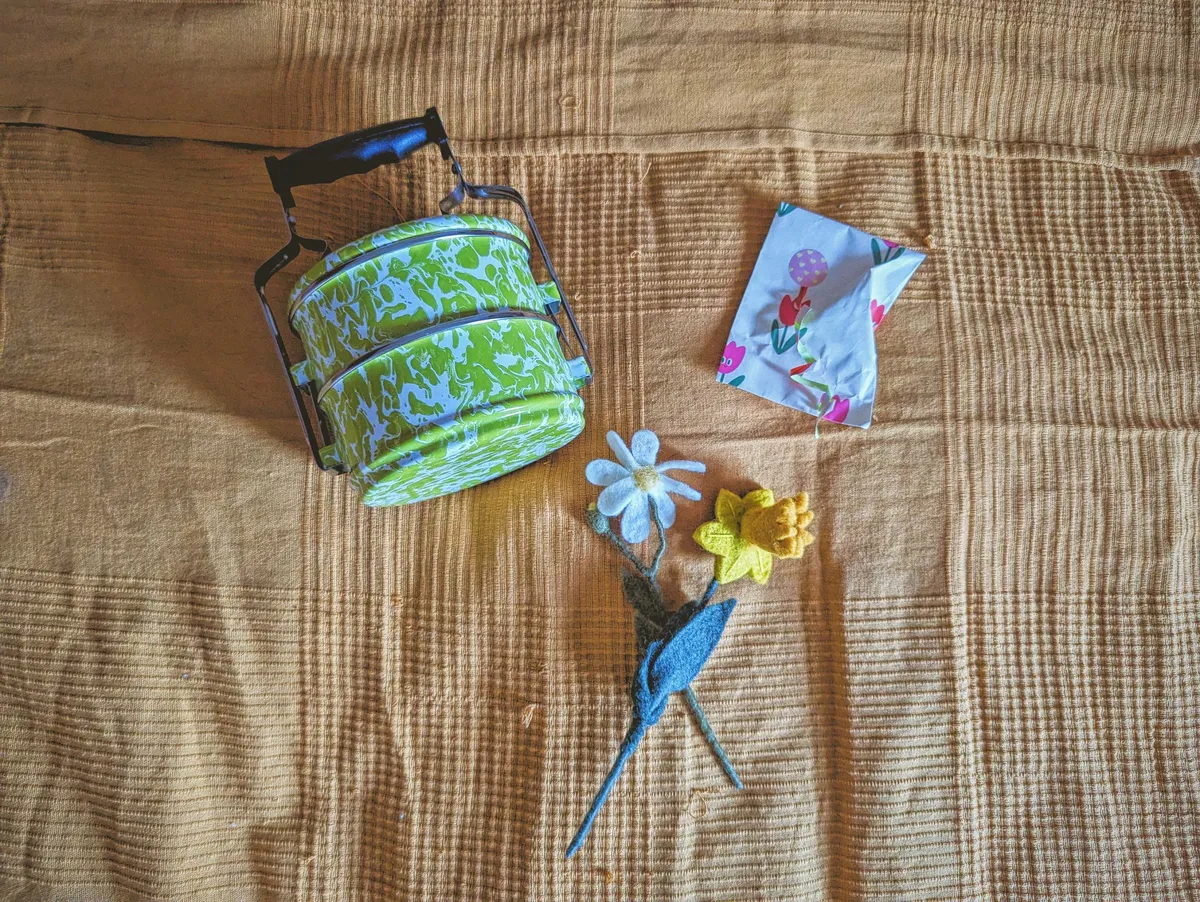 A green enamelware stacking lunchbox with handle, two felt flowers, and a small packaged gift.