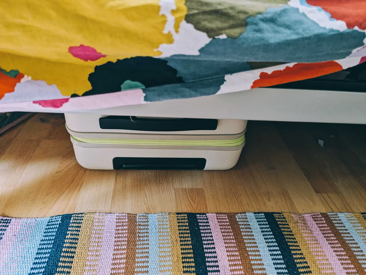 The beige suitcase is under the bed, surrounded by dust.