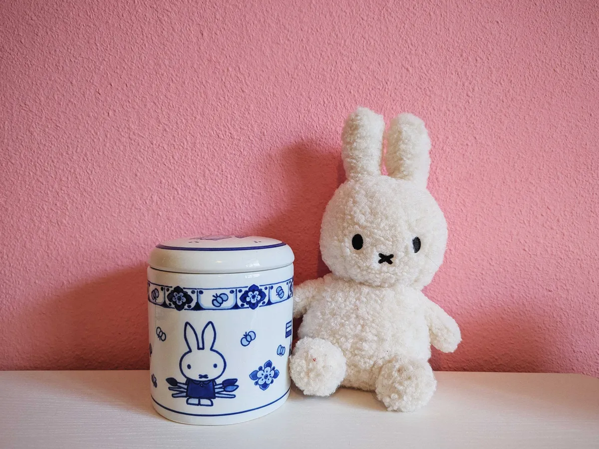 A blue and white Delfware Miffy jar, with a furry white Miffy plush doll next to it against a pink wall.