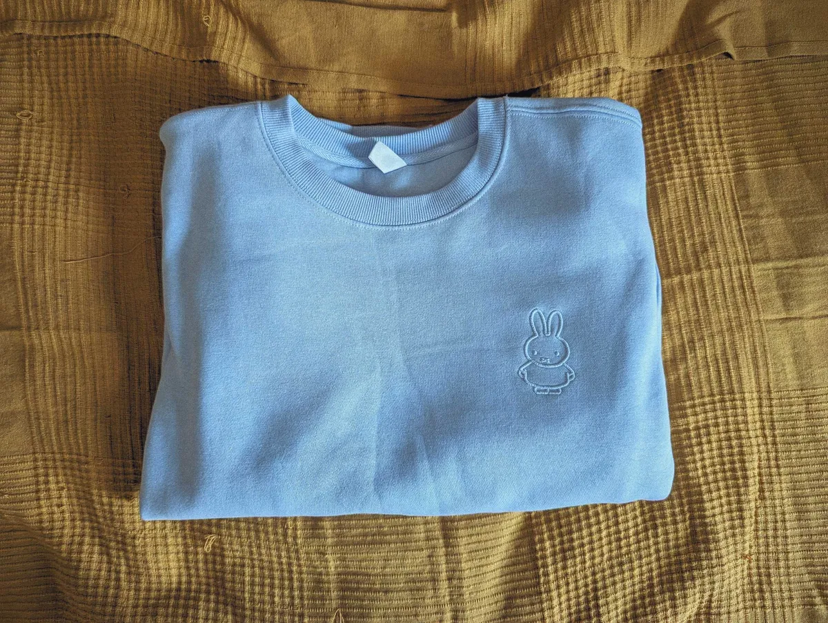 The front of a light blue sweater with a small embroidered Miffy on the left breast, in the same color.