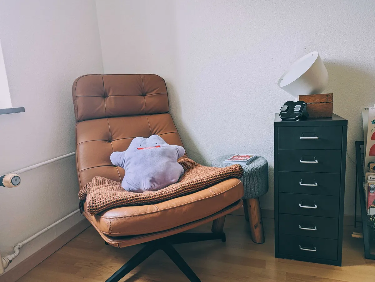 A clean corner with a brown leather lounge chair and a black metal drawer unit. There is a white Ikea PS 2016 on the drawer unit, along with a black View-Master and wooden box. A purple Ditto plush sits on the chair.