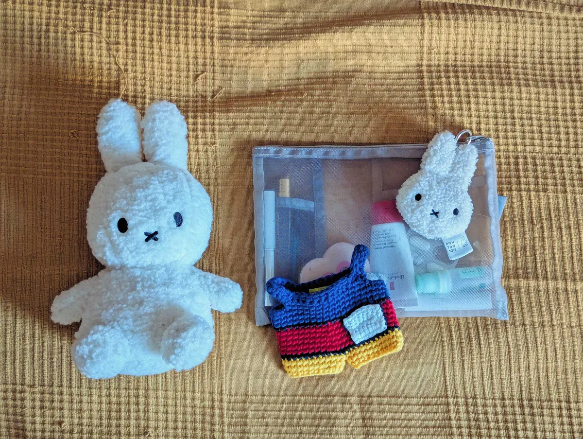 A white Miffy plush doll lies next to a mesh accessories pouch with a fluffy white Miffy keychain on the zipper. A crocheted blue, red, and yellow Mondrian-style doll-sized overall lies on top of the pouch.