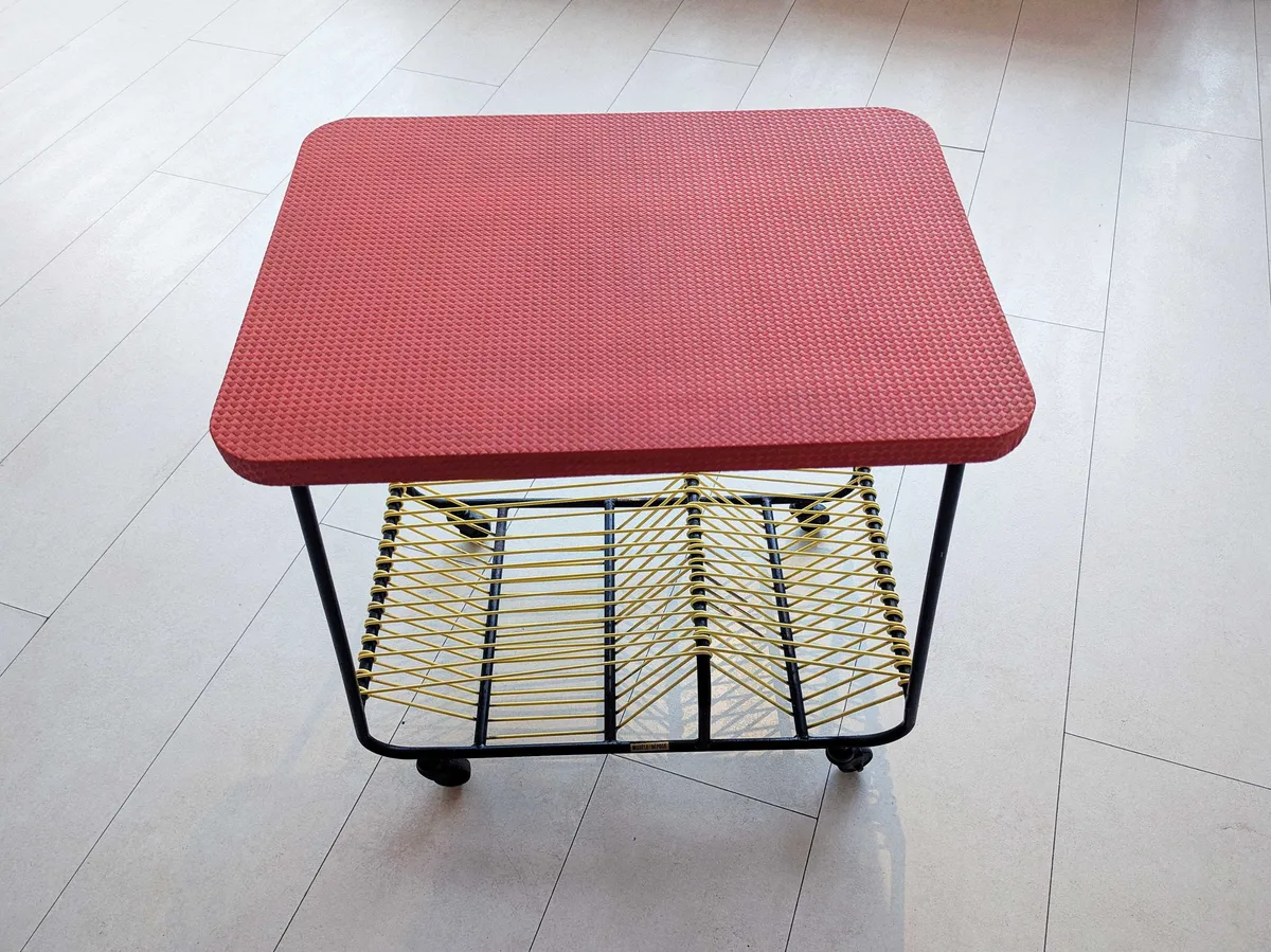 A table cart with a red rubber top, black metal rounded legs with wheels, and yellow rubber wire wrapped around the legs.