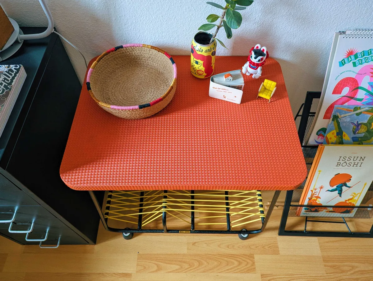Focusing on the red table top. There's an empty woven basket with accents of pink bronze, black, and red. Next to that is a planter made out of a beer can in yellow and red, a vinyl Japanese-style dog in white, red, and blue, a miniature yellow arm chair, a small white and red book, and a tiny ceramic sculpture of a white house with a red roof.