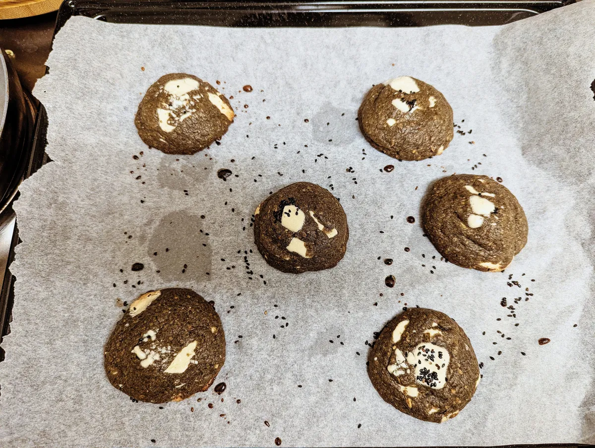 6 freshly baked cookies lay on top of a baking sheet. They are a dark gray color, filled with white chocolate chunks and sprinkled with black sesame seeds.