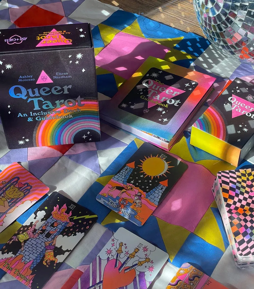A display of rainbow-colored tarot cards lie on a patterned quilt. A box reading, "Queer Tarot: An Inclusive Deck & Guidebook" stands upright.