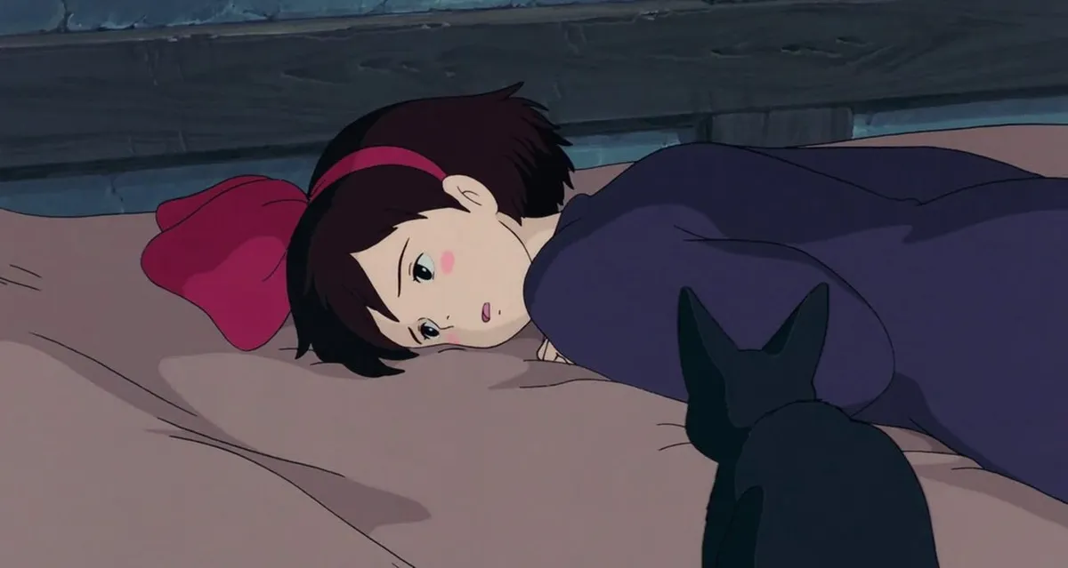 Kiki lying on the bed, looking mournful. Jiji stares at her, with her back to the viewer.