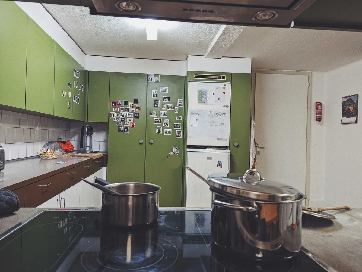 Two pots in the foreground on an electric stove. In the background is a kitchen with olive green cupboards. Polaroids are populated on some of the doors.