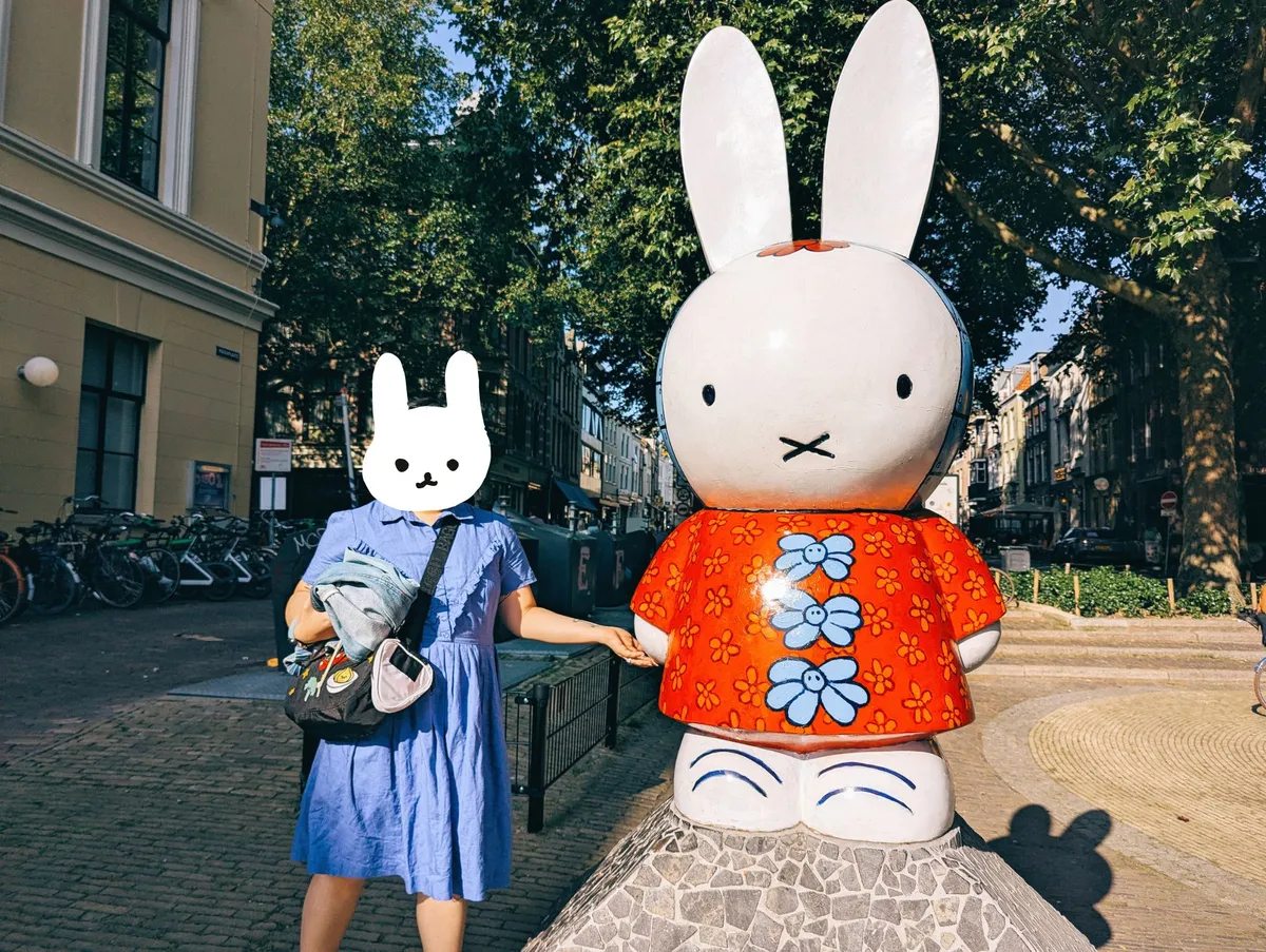 Peige, wearing a blue dress with their face hidden by a bunny illustration, poses next to a large Miffy statue. Miffy is wearing a red shirt with large blue flower buttons.