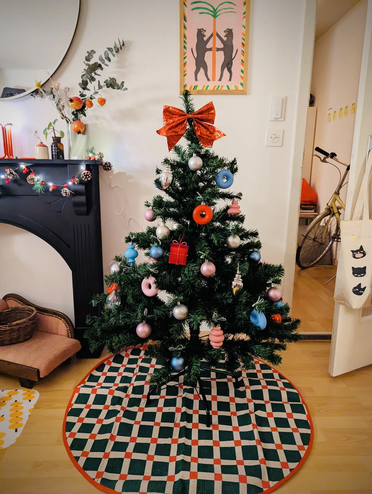 A green, plastic Christmas tree stands on top of a checkered circular tree skirt. The tree is decorated with red, pink, light blue, and silver ornaments. A big red bow is tied to the top of the tree.