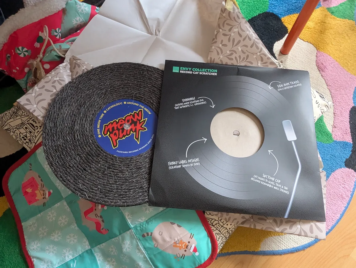 A cat scratcher shaped like a vinyl disk is laid on top of a bunch of items, such as wrapping paper and blankets. The "vinyl" has the label "Meow Punk" designed in the style of "Daft Punk."