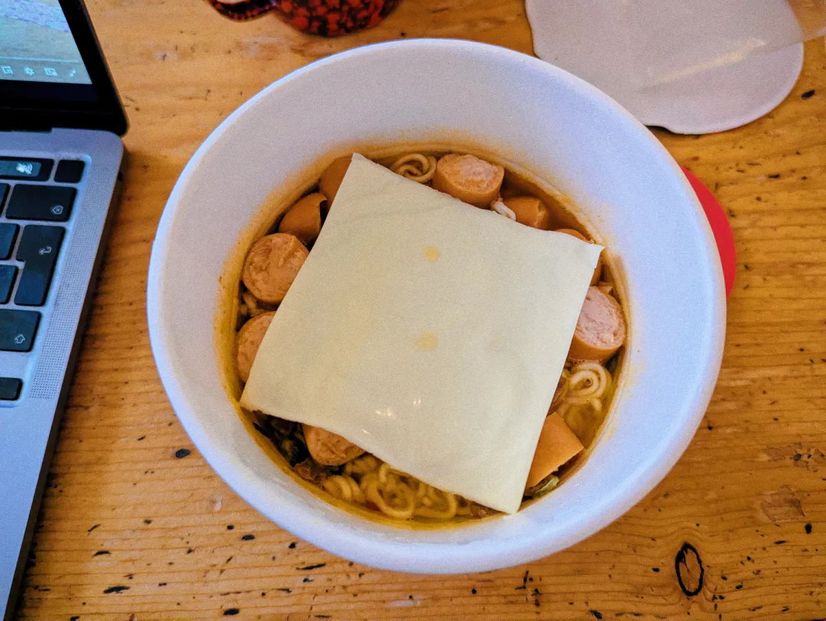 An overhead view of the instant ramen bowl. A square slice of white cheese covers the noodles, with bits of cut sausages peeking out.