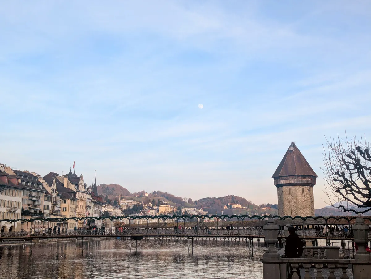 Lucerne’s Kappellbrucke with the mountains in the background, right before sunset. The moon is slight visible in the dusky blue sky.
