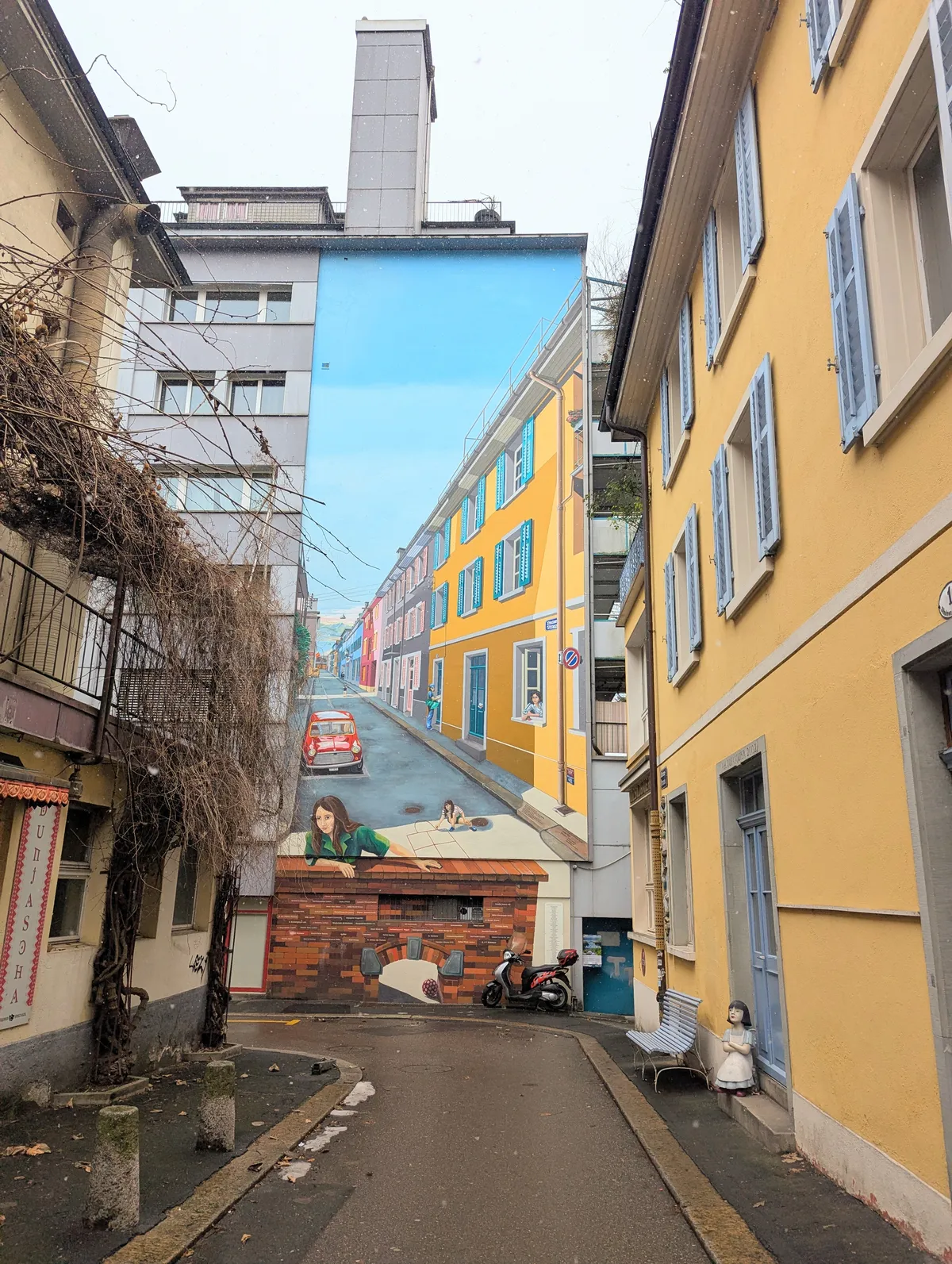 The end of a street with a mural facing the camera, which is a painting of the same street.
