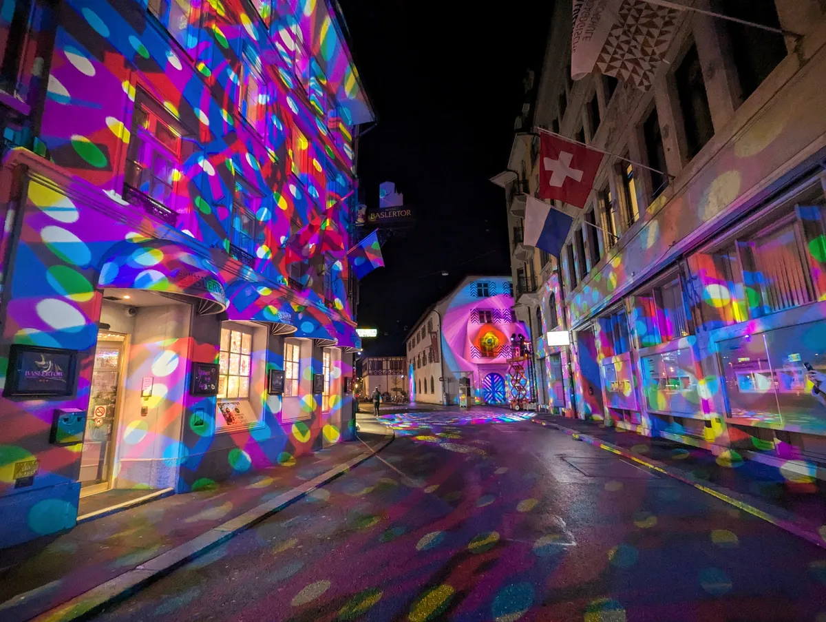 An empty, Swiss street at night, surrounded by old building fronts. The street and buildings are lit up with rainbow-colored circles projected on the walls and ground.