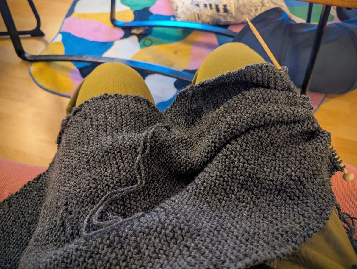 Dark grey knitted in-progress scarf draped across Peige's lap.