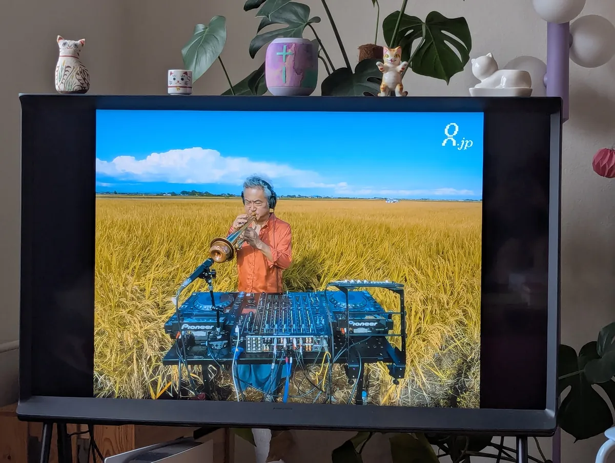 An older Japanese man with grey hair djing in a rice field. He's wearing headphones and is playing the trumpet directly into the microphone.