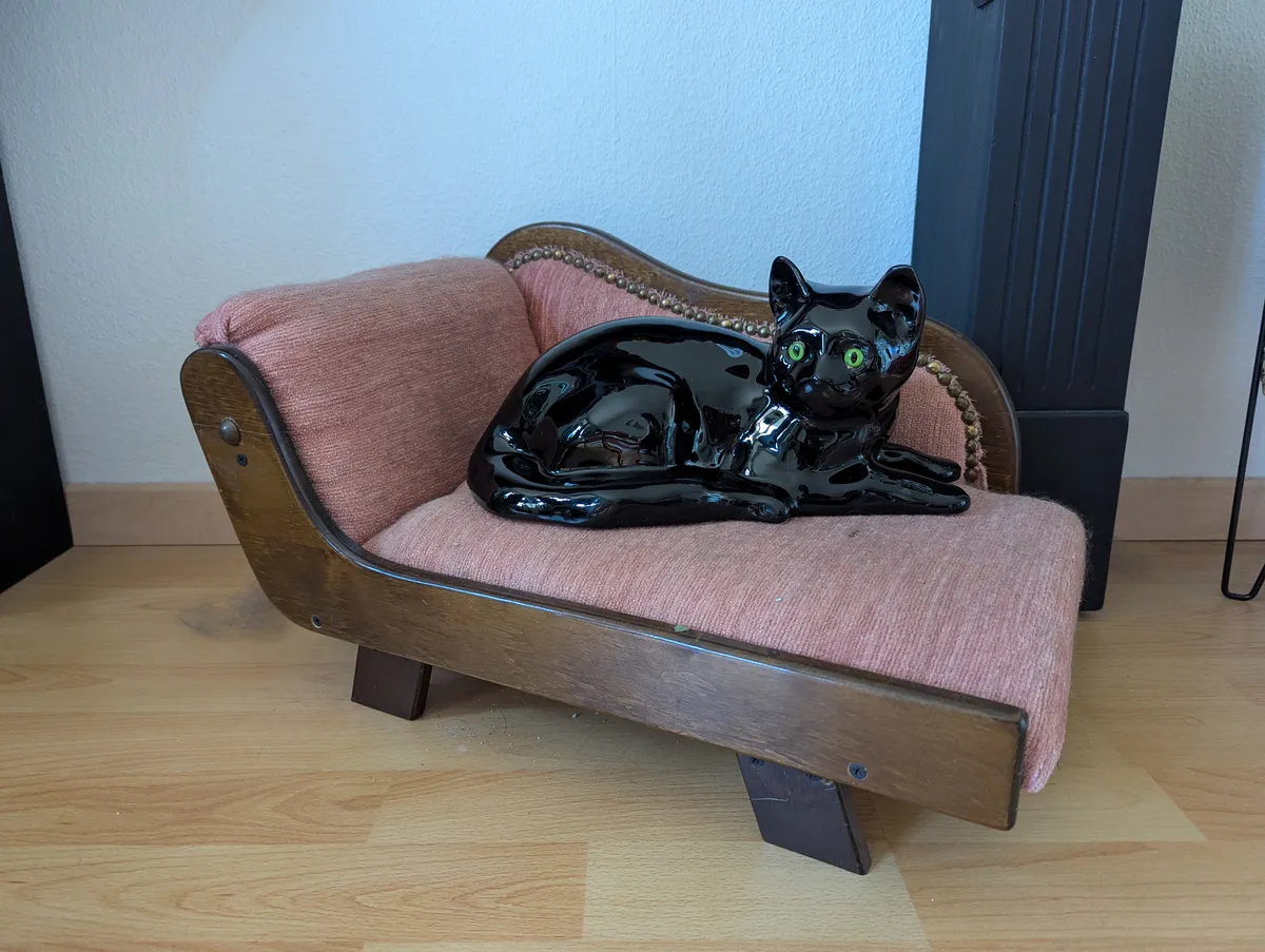 A realistic ceramic black cat with green eyes sits on a mini pink chaise lounge.
