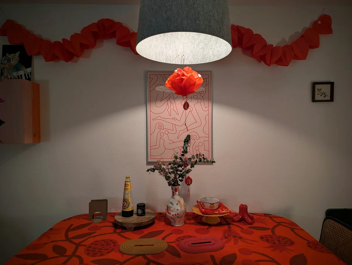 A dining table decorated with a red patterned table cloth and dried flower bouquet in the center. There is a red Chinese paper lantern hanging from the pendant lamp. There is a red, heart-patterned streamer hanging on the wall.