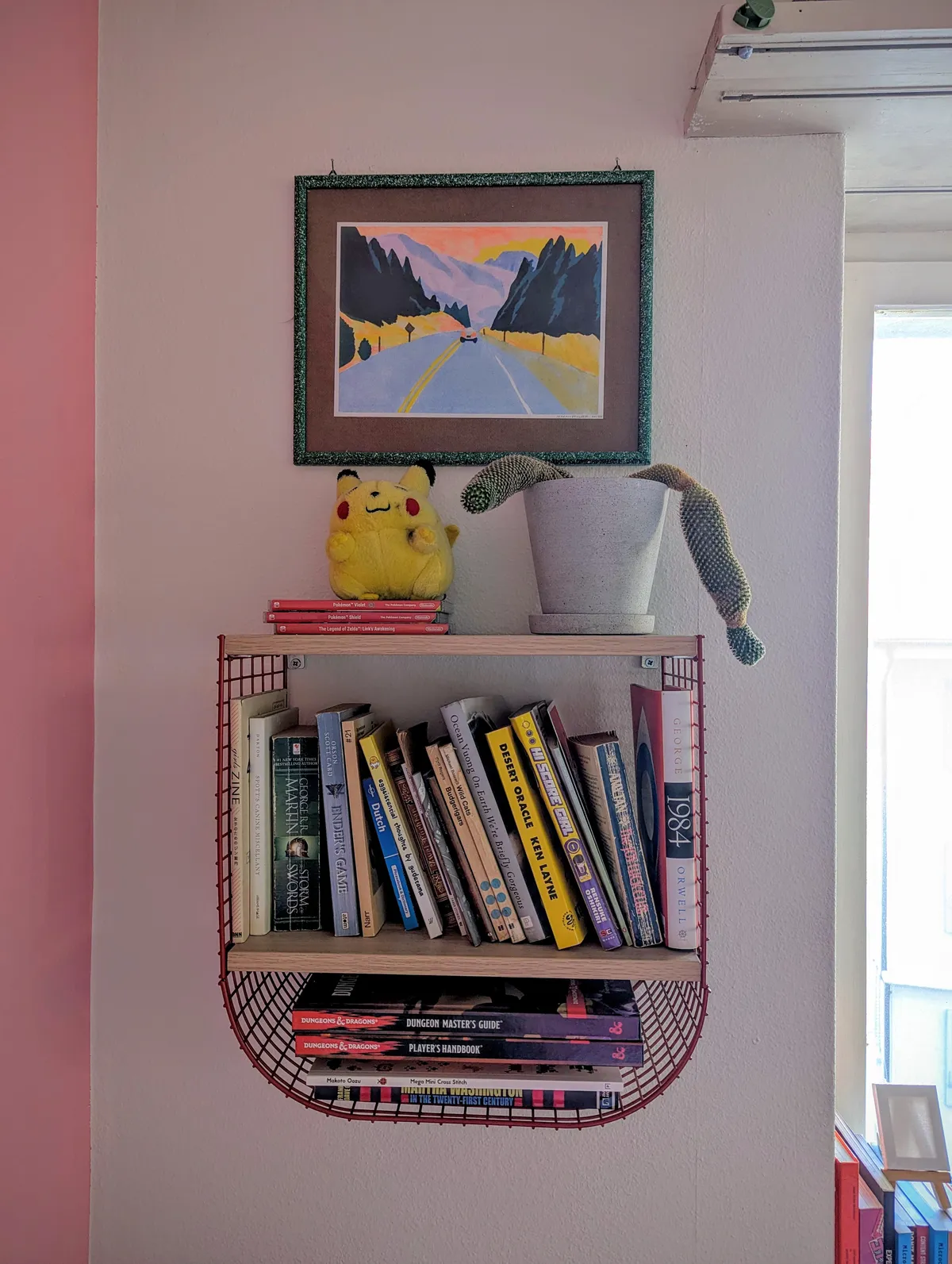 A risograph print of a car driving towards the mountains in a green frame hangs over a small bookshelf.