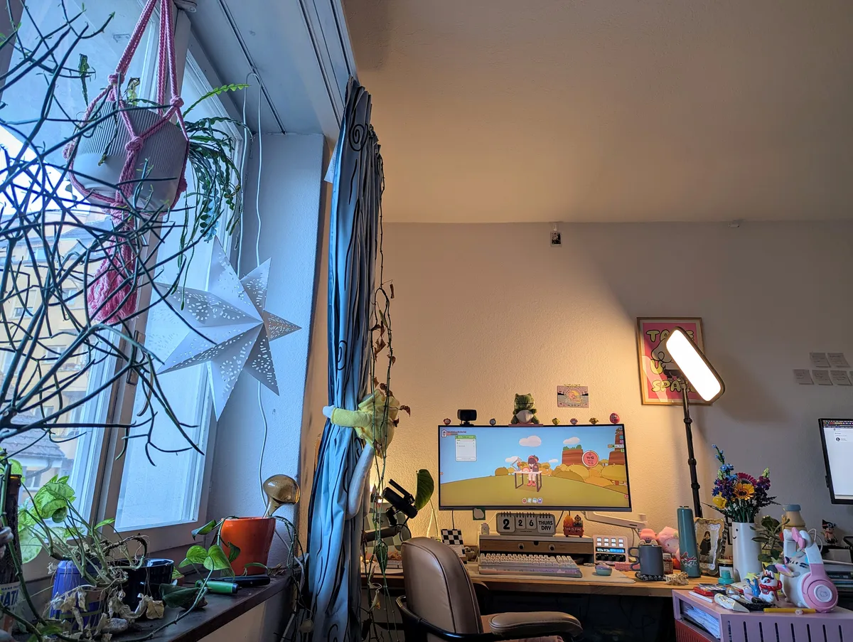 View of Peige's desk. To the left is the window with light streaming into the room. There are plants and a star lamp hanging from the window.