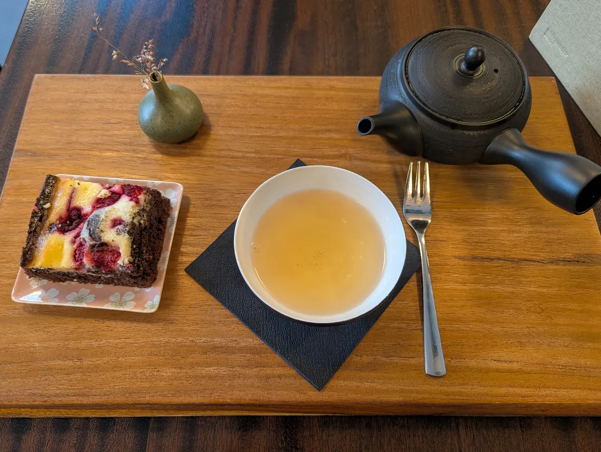 A wooden serving tray with a cup of tea and an ornate Chinese tea pot, and a square of brownie.