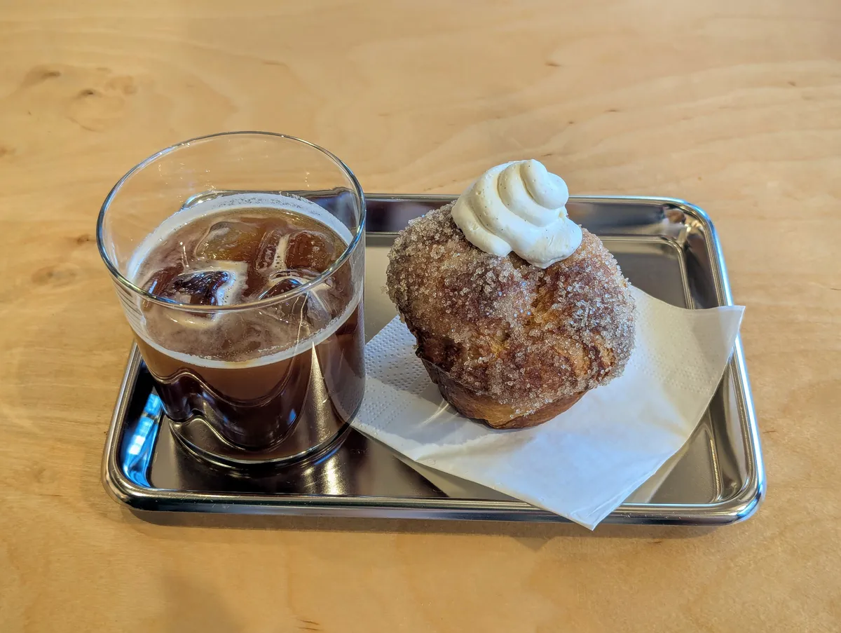 An iced Americano and a vanilla cream "cruffin" on a metal tray.