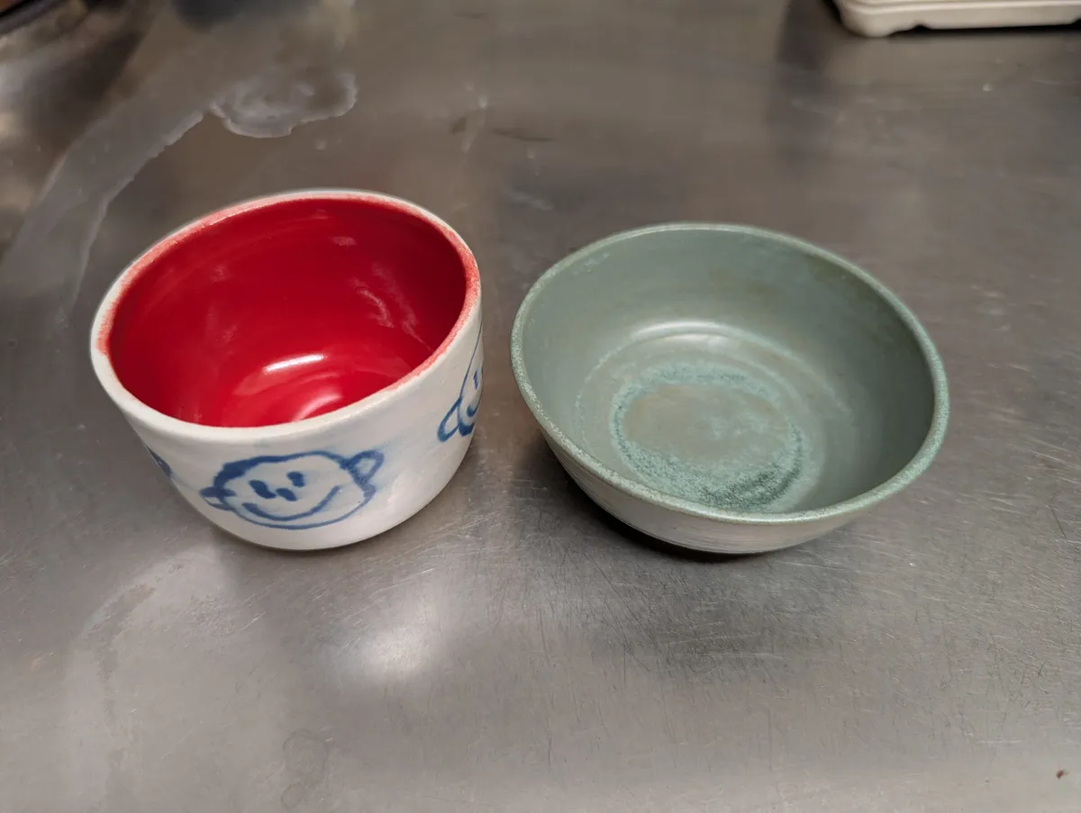 Two small ceramic cups, one with illustrations of happy faces and the other is a dark turquoise.