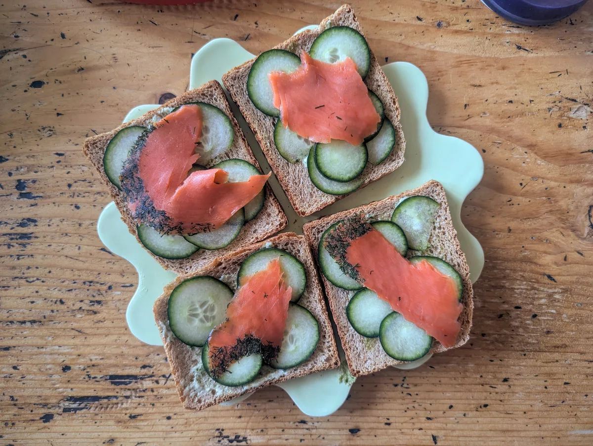 A light green squiggly plate with four slices of toast. Each toast has sliced cucumbers and a sliver of salmon.