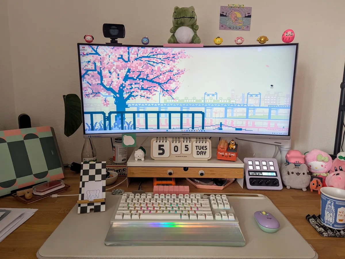 Peige's desk with a wide-screen monitor of a pixelate Sakura tree and lots of trinkets on the table. But it's organized.