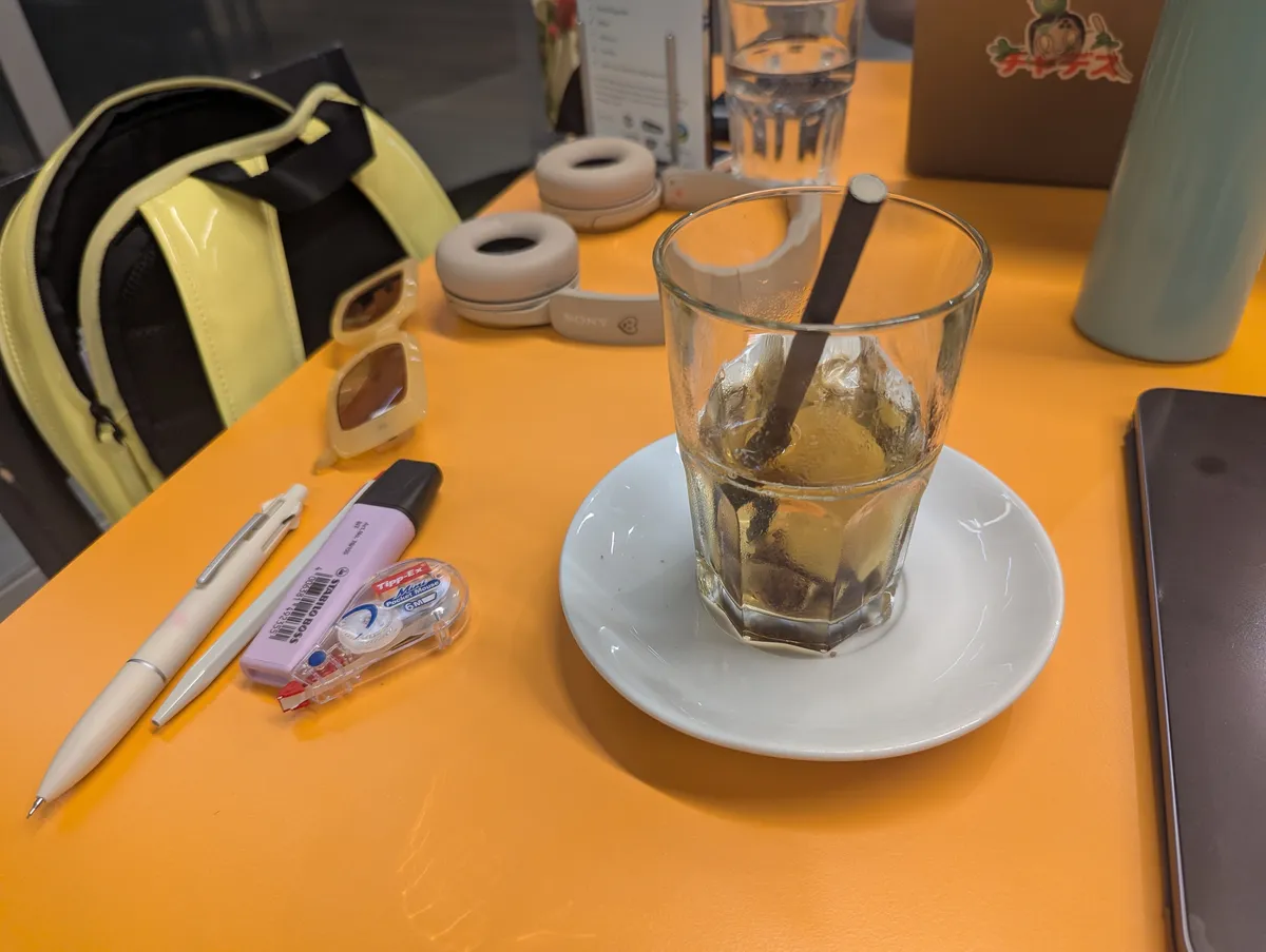 An orange table with a glass of iced tea, pens, and beige headphones.