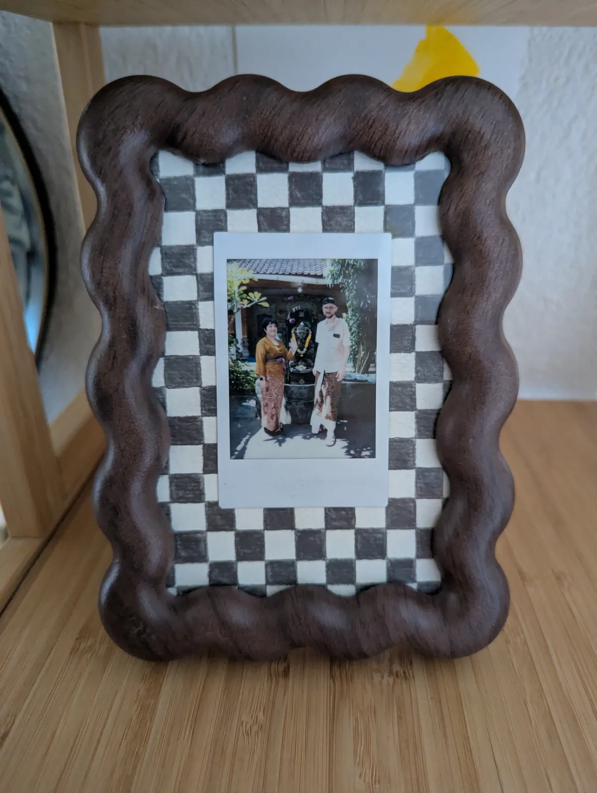 A framed fuji-film portrait of Peige and Daniel wearing traditional Balinese clothing. It is framed on a hand-painted checkered matte, and the frame is wavy and made of dark wood.